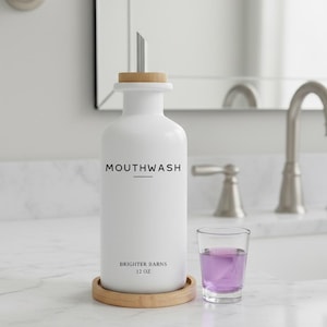 May include: A white mouthwash dispenser with a silver spout and a wooden lid, labeled "MOUTHWASH" and "BRIGHTER BARNS 12 OZ." It sits on a wooden coaster, with a glass of purple liquid beside it. The dispenser is 12 oz.