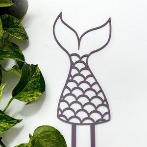 May include: Purple mermaid tail cake topper with a scale pattern. The topper has two prongs for inserting into a cake.