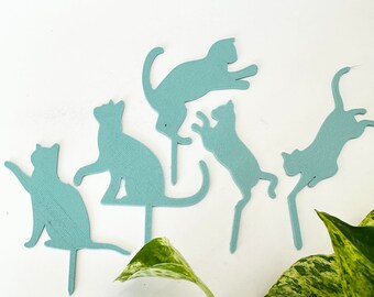 May include: Five teal blue cat silhouettes on sticks, arranged on a white surface with a green potted plant in the foreground.