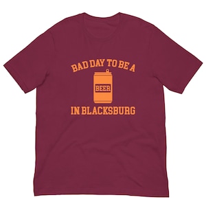 May include: Burgundy t-shirt with the text "BAD DAY TO BE A BEER IN BLACKSBURG" in orange. The design features an orange beer can graphic. A casual tee for beer lovers.