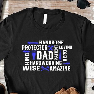 Op de afbeelding: Zwart T-shirt met een blauw en wit ontwerp met de tekst "Handsome, Protector, Patient, Loving, Dad, Hardworking, Wise, Amazing, Hero". Het ontwerp is gemaakt van gereedschap en andere symbolen.