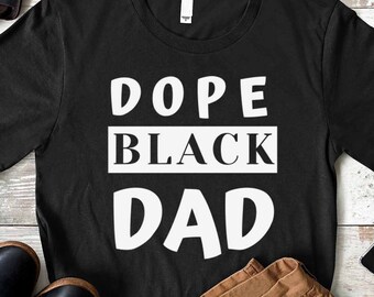 May include: Black t-shirt with white text that reads "Dope Black Dad". The shirt is laid out on a wooden surface with brown pants, a brown leather bag, sunglasses, and a phone.