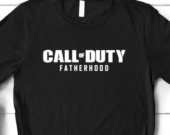 May include: Black t-shirt with white text that reads "Call of Duty Fatherhood". The shirt is laid flat on a white wooden surface with a brown leather bag, brown pants, and black shoes.