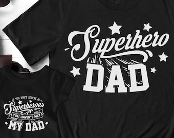 May include: Two black t-shirts with white text. The larger shirt says "Superhero Dad" with stars. The smaller shirt says "If you don't believe in superheroes you haven't met my dad".