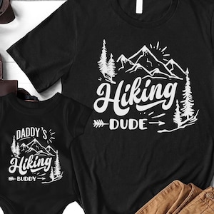 May include: Two black t-shirts with a white mountain and tree design. The larger shirt says "Hiking Dude" and the smaller shirt says "Daddy's Hiking Buddy".