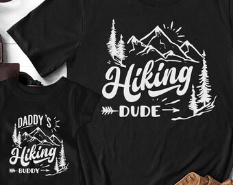 May include: Two black t-shirts with a white mountain and tree design. The larger shirt says "Hiking Dude" and the smaller shirt says "Daddy's Hiking Buddy".