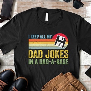 May include: A black t-shirt with a retro-style design featuring a floppy disk icon and the text "I keep all my dad jokes in a dad-a-base".