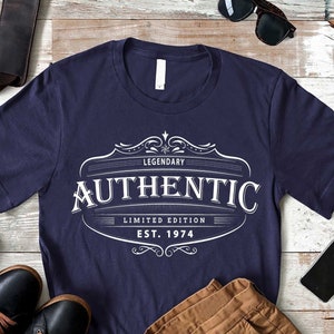 May include: A navy blue t-shirt with a white vintage-style logo that says "Legendary Authentic Limited Edition Est. 1974". The shirt is laid out on a white wood surface with brown pants, a brown leather bag, black shoes, sunglasses, and a cell phone.