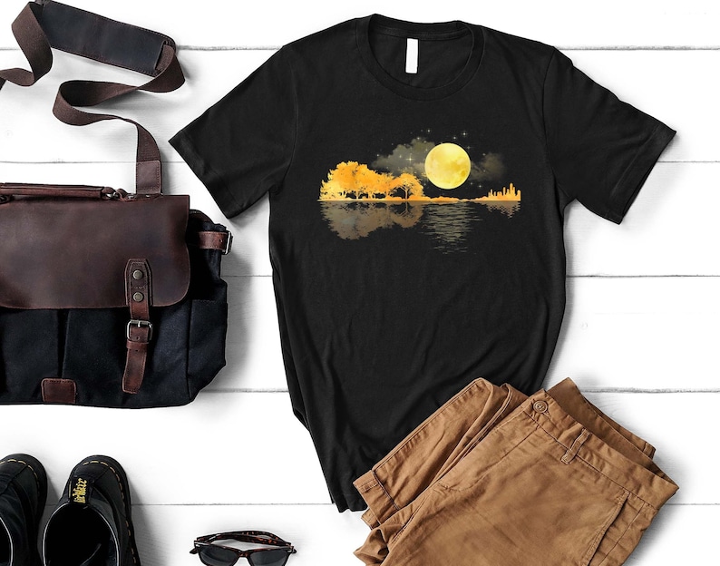 Puede incluir: Camiseta negra con un gr&aacute;fico de una silueta de guitarra con &aacute;rboles y una luna llena reflejada en el agua. La luna es amarilla y los &aacute;rboles son naranjas.