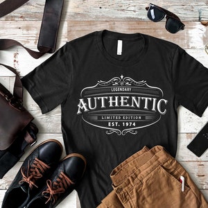 May include: Black t-shirt with a white vintage-style logo that says "Legendary Authentic Limited Edition Est. 1974". The shirt is laid out on a wooden surface with brown pants, a brown leather bag, sunglasses, and a cell phone.