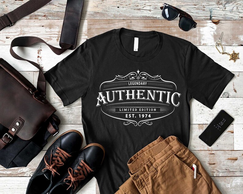 May include: Black t-shirt with a white vintage-style logo that says "Legendary Authentic Limited Edition Est. 1974". The shirt is laid out on a wooden surface with brown pants, a brown leather bag, sunglasses, and a cell phone.