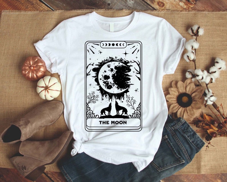 May include: White t-shirt with a black and white illustration of a tarot card depicting "The Moon". The card features a full moon with dripping black paint, two wolves, and a starry sky.