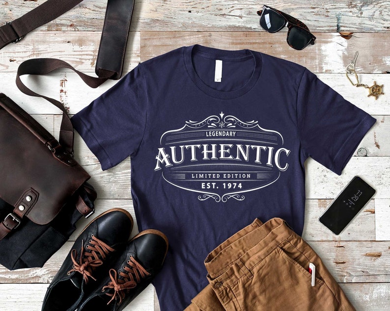 May include: A navy blue t-shirt with a white vintage-style logo that says "Legendary Authentic Limited Edition Est. 1974". The shirt is laid out on a white wood surface with brown pants, a brown leather bag, black shoes, sunglasses, and a cell phone.