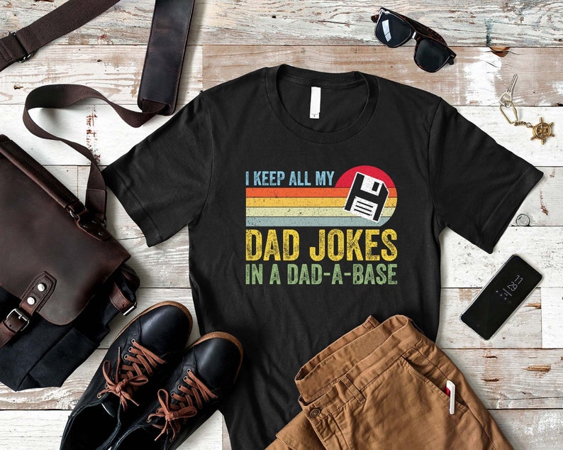 May include: A black t-shirt with a retro-style design featuring a floppy disk icon and the text "I keep all my dad jokes in a dad-a-base".