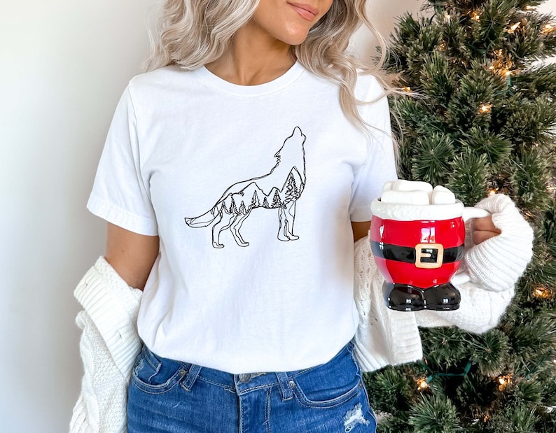 May include: White t-shirt featuring a black line drawing of a wolf howling at the moon. The wolf is formed from trees and mountains. The person wearing the shirt is holding a red Santa mug with marshmallows.