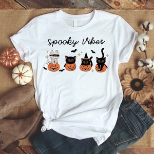 May include: White t-shirt with the text "Spooky Vibes" and a graphic of four black cats dressed in Halloween costumes sitting on pumpkins.