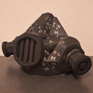 Könnte beinhalten: Eine schwarze Papiermaske mit geometrischem Design und weißen Buchstaben. Die Maske hat zwei runde Öffnungen an den Seiten und eine rechteckige Öffnung vorne mit einem schwarzen Gitter.