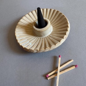 May include: A beige ceramic incense holder with a black incense cone. The holder has a ridged, oval design. Three wooden matches with pink tips are next to the holder on a gray surface.