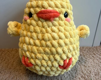 May include: A yellow crocheted chick plush toy with a red beak and black eyes. The chick has a fluffy texture and is sitting on a brown surface.