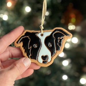May include: A ceramic ornament shaped like a Border Collie dog's head. The ornament features black and white glossy enamel with a tan border and a natural twine hanger. The dog's face is detailed with eyes and a nose.
