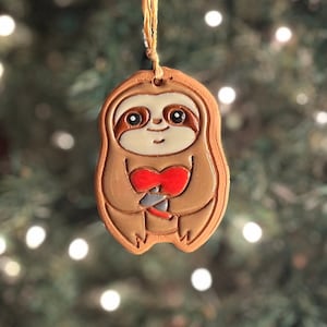 May include: A handmade ceramic sloth ornament with a heart-shaped design. The sloth is light brown with a white face, holding a red heart. It has a string for hanging. The background is a blurred Christmas tree.