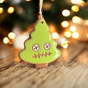 May include: A lime green Christmas tree ornament with stitched eyes and mouth, hanging from a string. The tree has a brown border and base. The background features blurred lights and a wooden surface.
