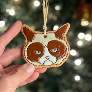 May include: A ceramic cat-shaped ornament with a grumpy expression. The ornament features brown, white, and light blue colors, with a beige border and a string for hanging. The cat's face is detailed with a frown and squinting eyes, perfect for a Christmas tree.