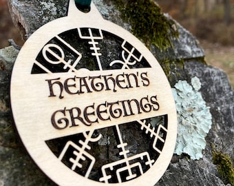 May include: A wooden ornament with a circular design and the text "Heathen's Greetings" in a decorative font. The ornament is hanging from a teal ribbon.