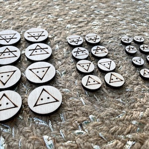 May include: A collection of small, round wooden tokens with various geometric symbols etched into them. The symbols include triangles and lines, with a dark brown outline. The tokens are arranged on a textured, neutral-toned surface.