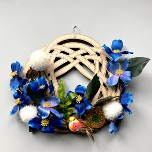 May include: A wooden wreath with a Celtic knot design, adorned with vibrant blue flowers, green leaves, and small white pom-poms. A decorative bird with red, yellow, and orange accents adds a touch of whimsy to this floral arrangement.