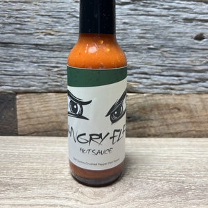 May include: A clear glass bottle of Angry Elf Hotsauce with a black cap. The label features a pair of angry eyes and the text "Angry Elf Hotsauce". The sauce is a vibrant red color.