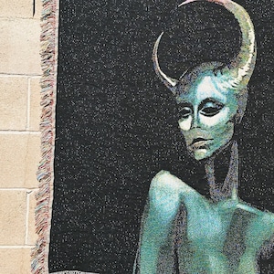 May include: A black woven tapestry with a fringed border features a green-toned figure with a crescent moon-shaped horn on their head. The figure has a neutral expression and is looking to the left.