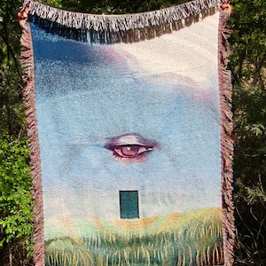 May include: A rectangular throw blanket with a surrealist design. The blanket features a large eye, a small green rectangle, and a landscape scene with a blue sky. The edges are fringed with a mix of colors.