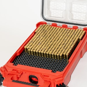May include: Open red plastic storage case with a clear lid. The case contains numerous small, cylindrical, brass-colored objects. The case has a black interior and is open, revealing its contents. The case is on a white background.