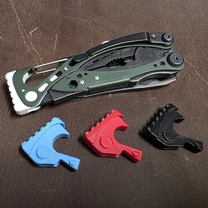 Op de afbeelding: Een groen en zwart multitool met een zilverkleurig mes en een witte clip. Drie gekleurde hulpstukken in blauw, rood en zwart liggen eronder.