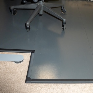 May include: A large, dark gray office chair mat with a black border. The mat is rectangular with a cut-out section. An office chair and a pet bed are on the mat, which sits on a carpeted floor.