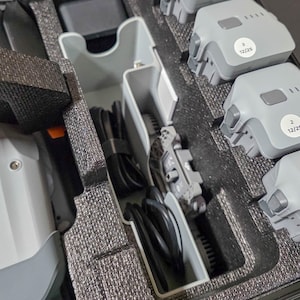 May include: A grey drone and its accessories are neatly arranged inside a black carrying case. The case includes the drone, propellers, batteries, charging cables, and other components. The batteries are labelled with the numbers 2, 3, and 4.