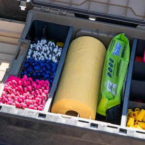 May include: A tan plastic storage case containing organized compartments. The left section holds rows of pink, blue, and white looped fabric strips. The center contains a large yellow paper roll and a green package of Fierce disinfectant wipes. The right section holds pink tape rolls and small yellow plastic cones.