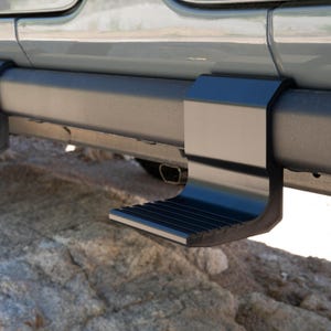 May include: A matte black metal side step accessory mounted onto the horizontal frame rail of a vehicle. The step features a flat, textured horizontal platform with a vertical support bracket that attaches securely to the vehicle's chassis.