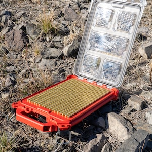 Puede incluir: Maleta roja abierta con tapa transparente, llena de numerosos objetos peque&ntilde;os de color lat&oacute;n. La maleta descansa sobre una superficie rocosa al aire libre. La tapa tiene m&uacute;ltiples compartimentos y el texto "Milwaukee Packout" es visible.