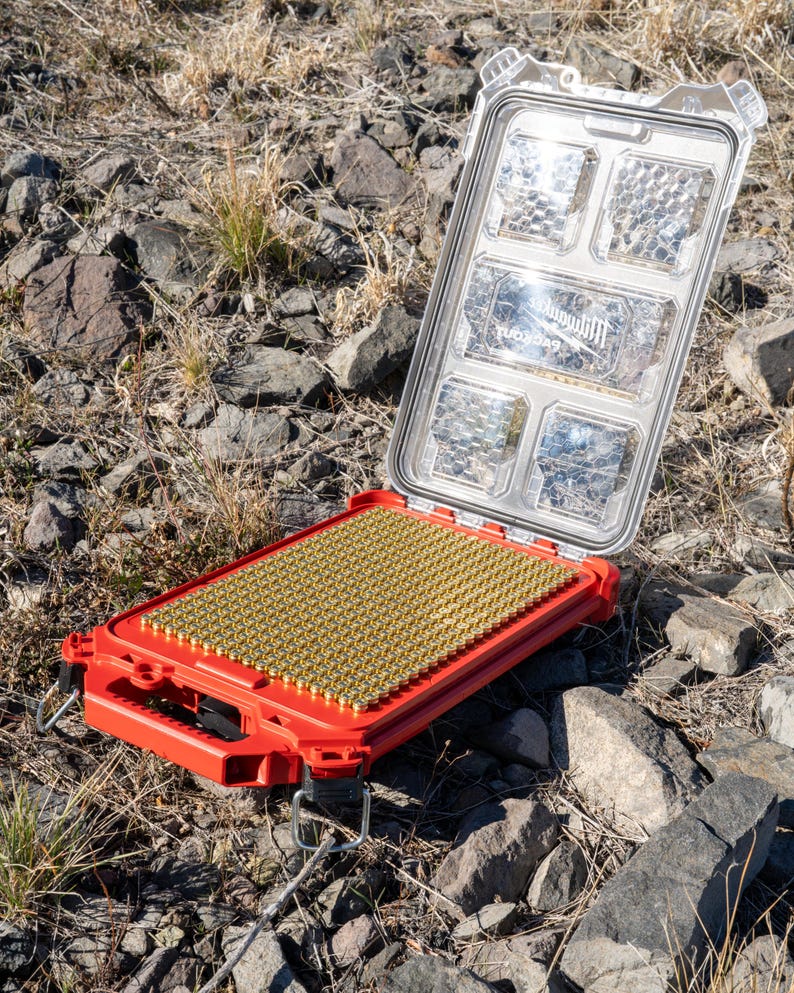 Puede incluir: Maleta roja abierta con tapa transparente, llena de numerosos objetos peque&ntilde;os de color lat&oacute;n. La maleta descansa sobre una superficie rocosa al aire libre. La tapa tiene m&uacute;ltiples compartimentos y el texto "Milwaukee Packout" es visible.
