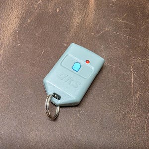 May include: A light gray DKS remote control with a silver key ring. The remote has a blue button, a red indicator light, and the letters "DKS" printed on the front. The remote is rectangular and has rounded edges.