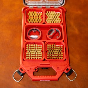 May include: Open red ammunition storage case with a clear lid. The case is divided into compartments labelled with ammunition types: .45 ACP, .40 S&W, 9 mm, and .380 Auto. The word "Ammo" is printed on the handle.