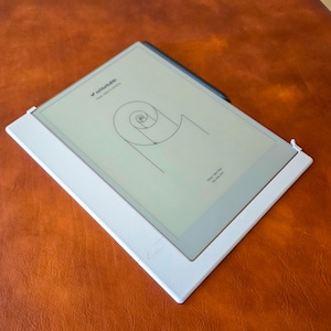 May include: A white tablet with a black stylus, showing a spiral design. The tablet is in a white case, placed on a brown surface. The screen displays the word "remarkable" at the top.
