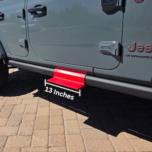 May include: A bright red metal step accessory attached to the side of a gray Jeep Wrangler vehicle. The step features a flat horizontal surface and a vertical support, with a white graphic label indicating a length of 13 inches.