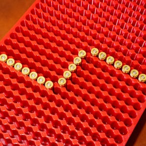 Puede incluir: Una bandeja roja de pl&aacute;stico para munici&oacute;n llena de balas con casquillos de lat&oacute;n. Las balas est&aacute;n dispuestas en zigzag. La bandeja tiene un dise&ntilde;o de panal con numerosas peque&ntilde;as hendiduras circulares para sujetar las balas.