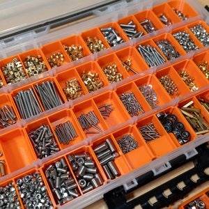 May include: A clear plastic organizer with orange compartments filled with various metal hardware. The hardware includes screws, bolts, nuts, and other small parts in silver, gold, and brass tones. The organizer is open, revealing the contents.