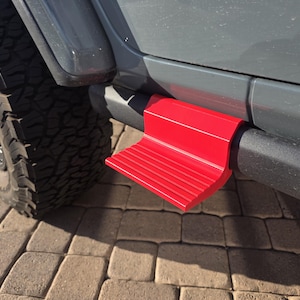 May include: A bright red, L-shaped vehicle step accessory attached to the black side rail of a gray vehicle. The step features a textured, ribbed surface for traction and is mounted directly onto the tubular frame near the front tire.