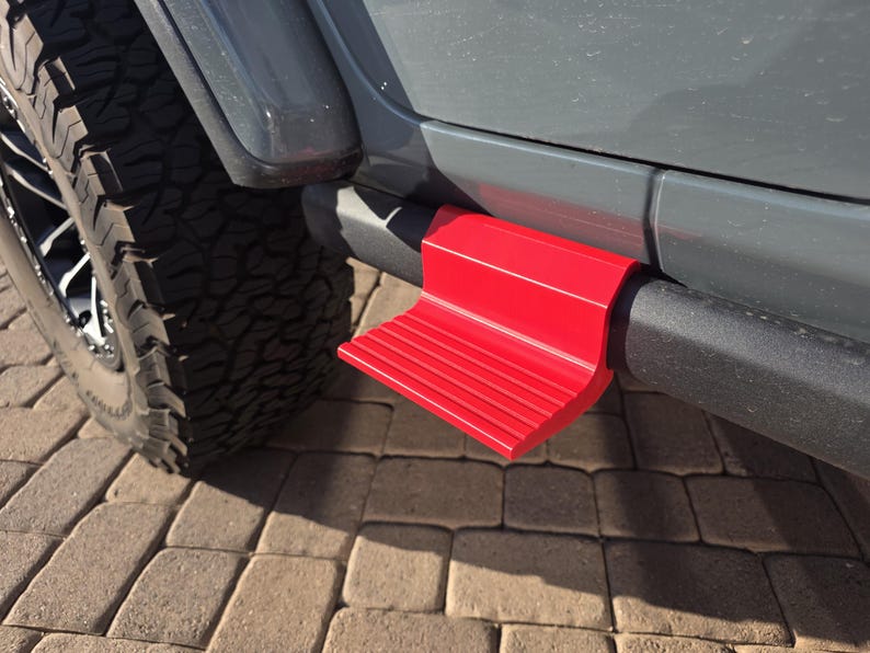 May include: A bright red, L-shaped vehicle step accessory attached to the black side rail of a gray vehicle. The step features a textured, ribbed surface for traction and is mounted directly onto the tubular frame near the front tire.