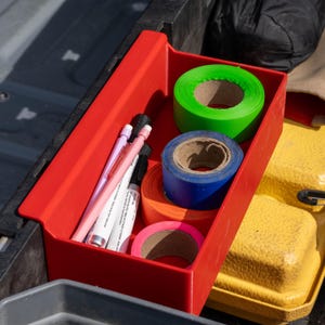 May include: A rectangular red plastic storage bin containing three pens and four rolls of tape. The tape rolls are neon green, blue, orange, and pink. The pens include one light pink pen and two white pens with black text and black caps.